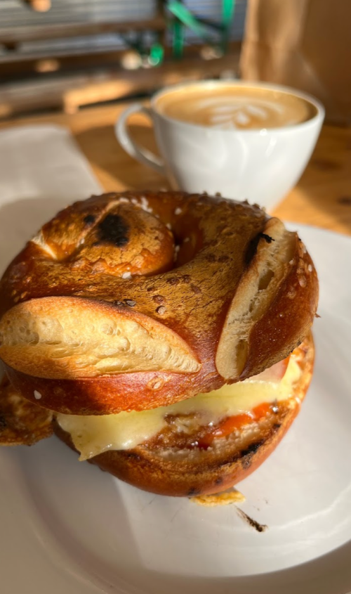 A toasted pretzel sandwich with cheese and tomato, accompanied by a cup of coffee in the background.
