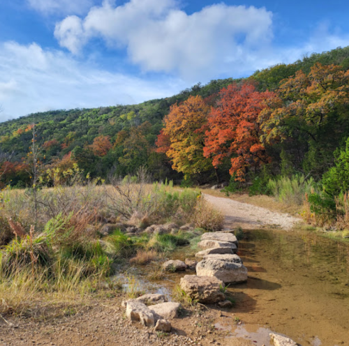 A scenic pathway with colorful autumn trees and a small stream, surrounded by lush greenery under a blue sky.
