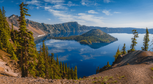 A serene view of Crater Lake, surrounded by tall trees and mountains, reflecting a clear blue sky.