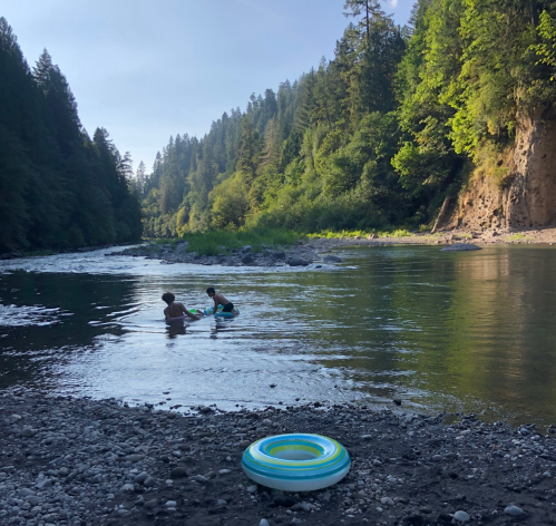 Two children play in a river surrounded by tall trees and rocky banks, with a colorful inner tube in the foreground.