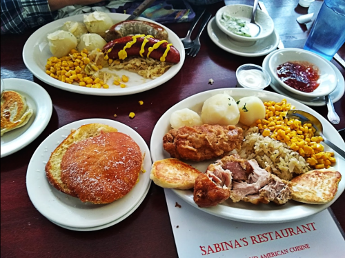 A table with two plates of food: one with fried chicken, potatoes, corn, and rice; the other with sausages and pancakes.