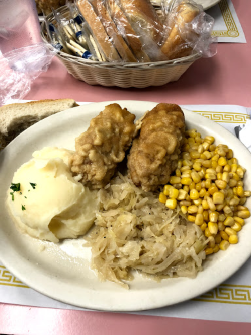 A plate of food featuring fried chicken, mashed potatoes, corn, and sauerkraut, with bread rolls in the background.