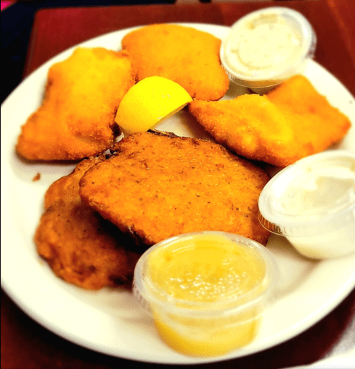 A plate of golden fried fish fillets with lemon and dipping sauces on the side.
