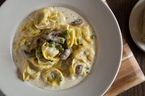 A bowl of creamy pasta with yellow tortellini, mushrooms, and green peas, garnished with black pepper.