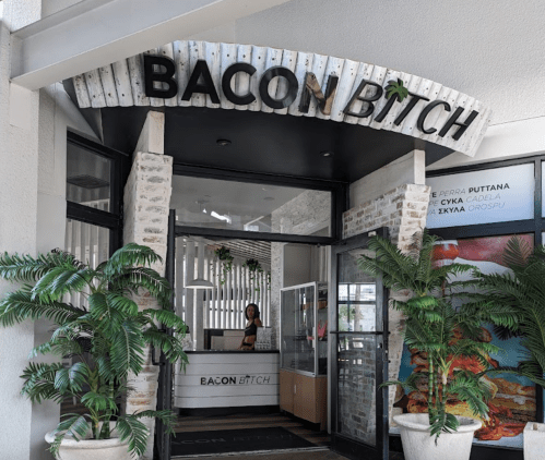 Entrance of a restaurant named "Bacon B*tch," featuring a modern design and greenery. A staff member is at the counter.