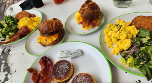 A table with four plates of breakfast: scrambled eggs, bacon, croissant sandwich, hash browns, and pancakes with syrup.