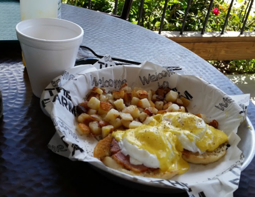 A plate of eggs Benedict with ham and hollandaise sauce, served with seasoned potato cubes and a cup of drink.