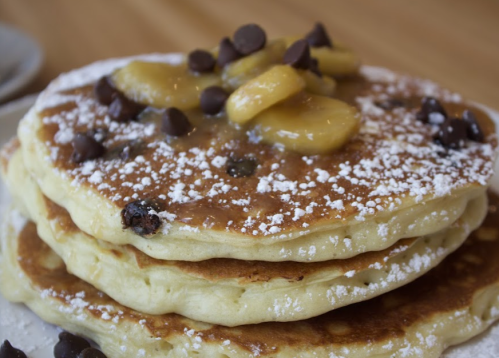 Stack of fluffy pancakes topped with banana slices, chocolate chips, and a dusting of powdered sugar.