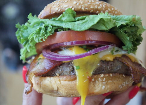 A close-up of a juicy burger with lettuce, tomato, onion, bacon, and melted cheese, held in a hand.