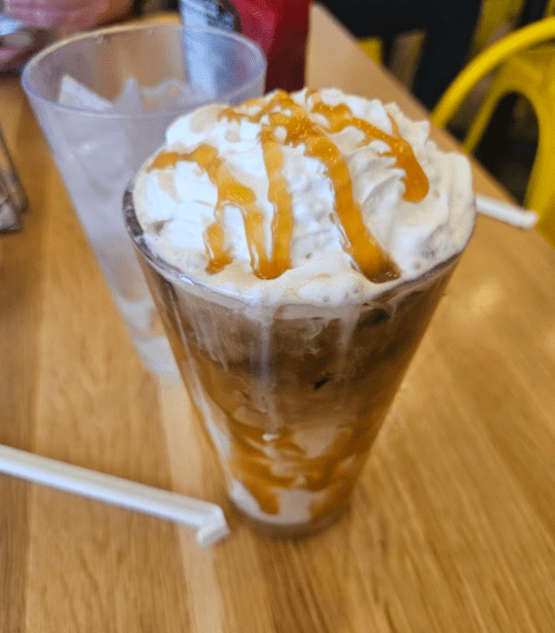 A tall glass filled with iced coffee topped with whipped cream and drizzled with caramel sauce, on a wooden table.