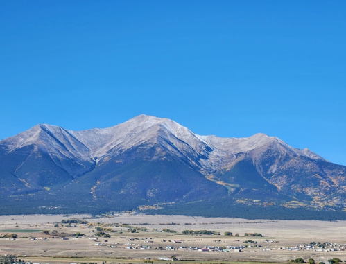 A panoramic view of majestic mountains under a clear blue sky, with a valley and small town visible below.