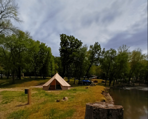 A tent by a calm river, surrounded by green trees and a cloudy sky, with picnic areas in the background.