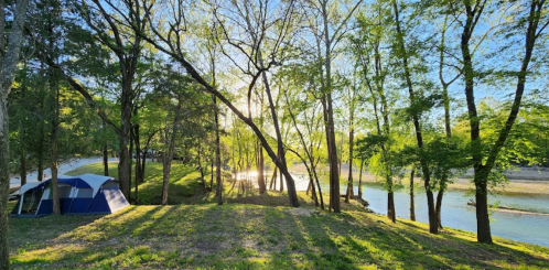 A serene campsite by a river, surrounded by trees and sunlight filtering through the leaves.