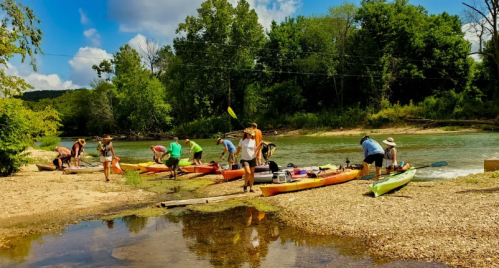 Group of people preparing kayaks by a riverbank surrounded by trees and a blue sky with clouds.