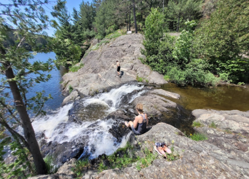 Two people relax by a rocky riverbank with a small waterfall, surrounded by trees and greenery.