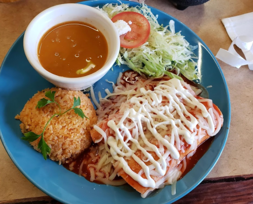 A blue plate with enchiladas drizzled in sauce, rice, salad, and a small bowl of soup.