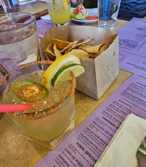 A refreshing cocktail with lime and lemon slices, served with a side of tortilla chips in a paper container.