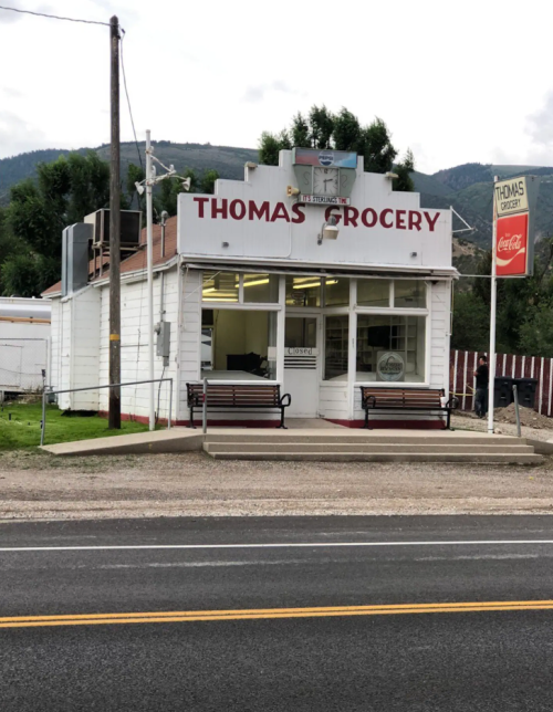 A small, white grocery store with a sign reading "Thomas Grocery" and benches outside, set against a mountainous backdrop.