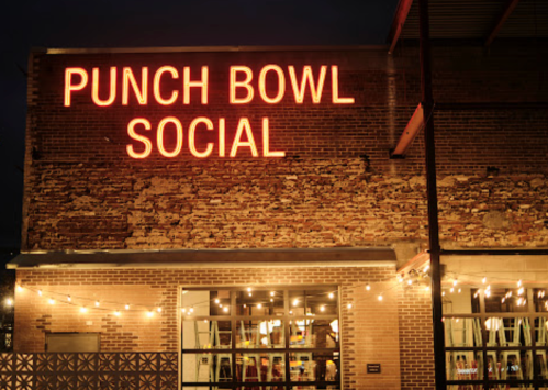 Neon sign reading "PUNCH BOWL SOCIAL" on a brick building, illuminated at night with string lights in the foreground.