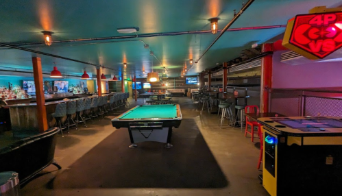 A spacious bar with a pool table, arcade games, and seating areas, featuring dim lighting and a vibrant atmosphere.