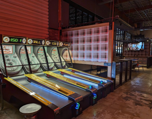 A dimly lit arcade with three skee ball machines and a bar area in the background.