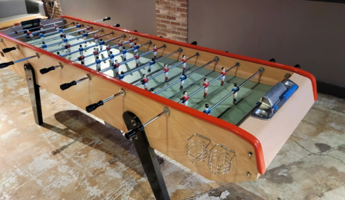 A foosball table with red and blue players, set in a casual indoor space with exposed brick walls.