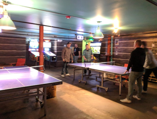 A group of people playing ping pong in a casual game room with arcade machines in the background.