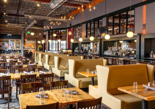 Interior of a modern restaurant featuring wooden tables, yellow booths, and a bar area with pendant lighting.