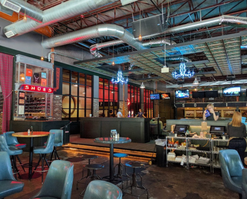 A modern bar interior with neon signs, high tables, and a lively atmosphere, featuring a bartender and patrons.