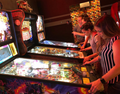 Four people playing pinball machines in a vibrant arcade setting.