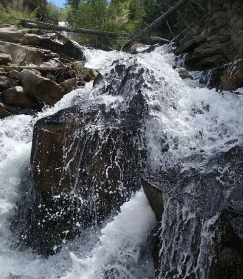 A cascading waterfall flows over large rocks, surrounded by lush greenery and natural scenery.