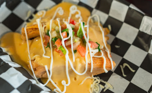 A plate of tamales topped with cheese sauce, diced tomatoes, lettuce, and drizzled with sour cream on a checkered paper.