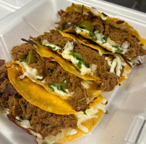 A close-up of four tacos filled with shredded beef, onions, cilantro, and jalapeños in a takeout container.
