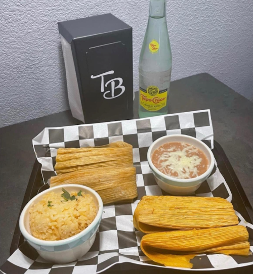 A tray with tamales, rice, beans topped with cheese, and a bottle of Topo Chico, all on a checkered paper.