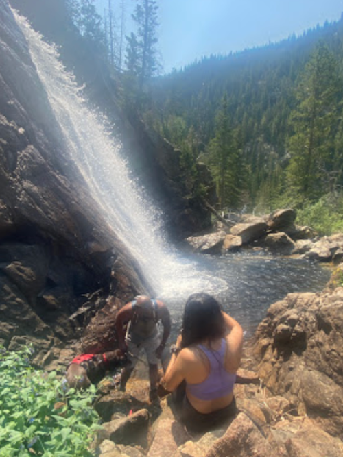 A waterfall cascades down rocky terrain, with two people near the water's edge surrounded by trees and nature.