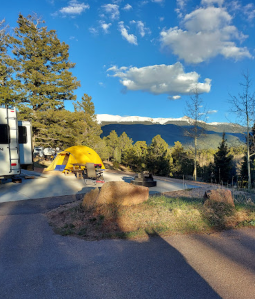 A yellow tent and RV are set up in a scenic campsite with mountains and trees under a blue sky.
