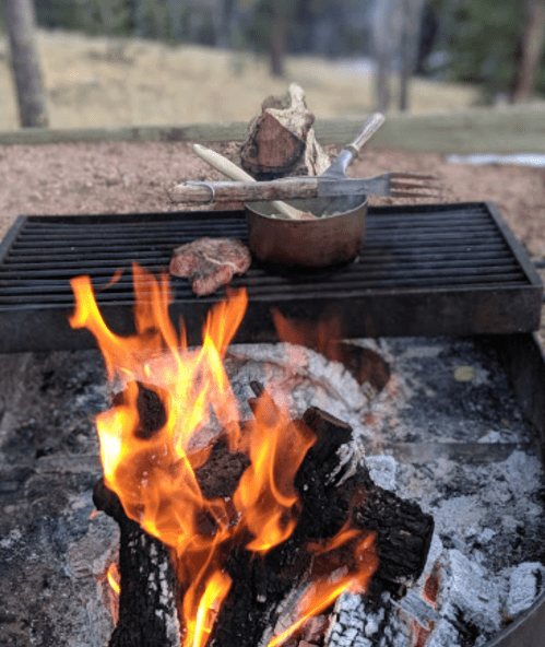 A campfire with flames, a grill, cooking utensils, and a pot, set in a natural outdoor environment.