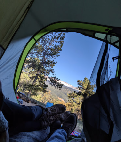 View from inside a tent, showing two pairs of feet and a scenic landscape with trees and mountains outside.