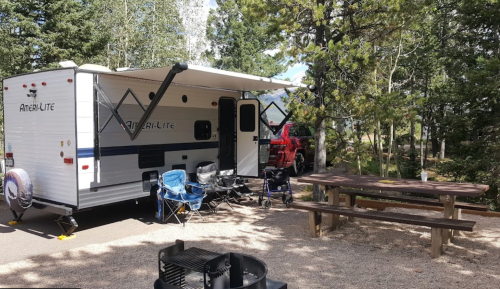 A travel trailer parked in a wooded area, with camping chairs and a picnic table nearby.