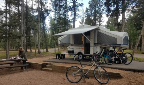 A camper trailer in a wooded area, with a person at a picnic table and bicycles nearby.