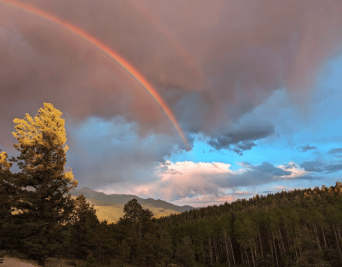 A vibrant rainbow arcs over a mountainous landscape, framed by dark clouds and lush green trees.