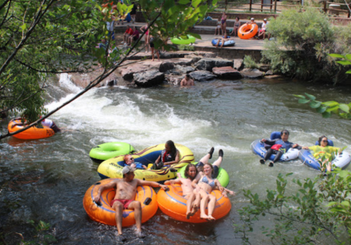People enjoying a sunny day floating on colorful inner tubes in a river, surrounded by greenery and rocks.