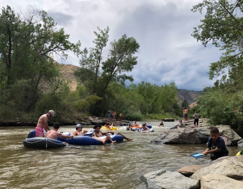 People enjoying a river day, floating on tubes and relaxing on rocks surrounded by trees and a cloudy sky.