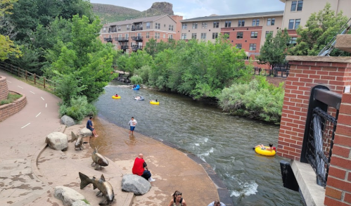 A scenic river with people kayaking and relaxing by the shore, surrounded by greenery and buildings in the background.