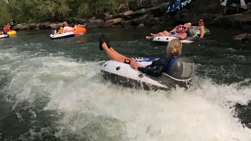 A group of people relaxing on inflatable tubes in a river, with splashing water and rocky banks in the background.