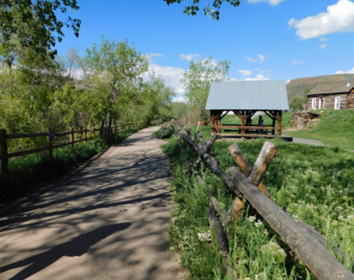 A scenic pathway lined with trees and a wooden fence, leading to a gazebo under a blue sky with fluffy clouds.