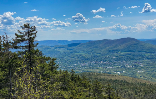 A panoramic view of rolling green mountains and a small town under a blue sky with fluffy clouds.