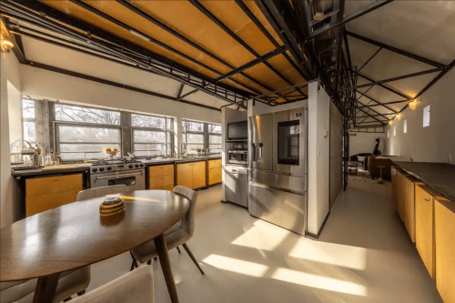 Modern kitchen with stainless steel appliances, wooden cabinets, and a round dining table, featuring large windows for natural light.