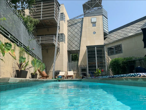 A clear swimming pool in the foreground, with a modern building and greenery in the background under a blue sky.