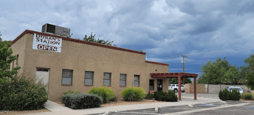 A tan building with a sign reading "Duran's Station - Open" under a cloudy sky, surrounded by greenery and a parking lot.